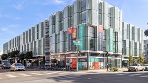 En moderne bygning med store glasfacader, et tydeligt skilt med ’Sælges nu’ og et gadebillede med parkerede biler og cykler.