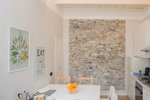 Un área de comedor con pared de piedra, una mesa blanca y sillas. Hay cuadros en la pared y un cuenco de fruta sobre la mesa.