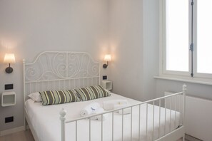Ein weißes Metallbett mit ornamentalem Kopfteil, zwei Nachttischlampen, ein Fenster mit Ausblick und ein kleiner Nachttisch mit Lampe.
