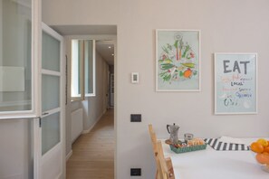 Un pasillo con piso de madera, una puerta blanca y una ventana. Hay dos cuadros en la pared, uno con verduras y otro con texto sobre la comida local.
