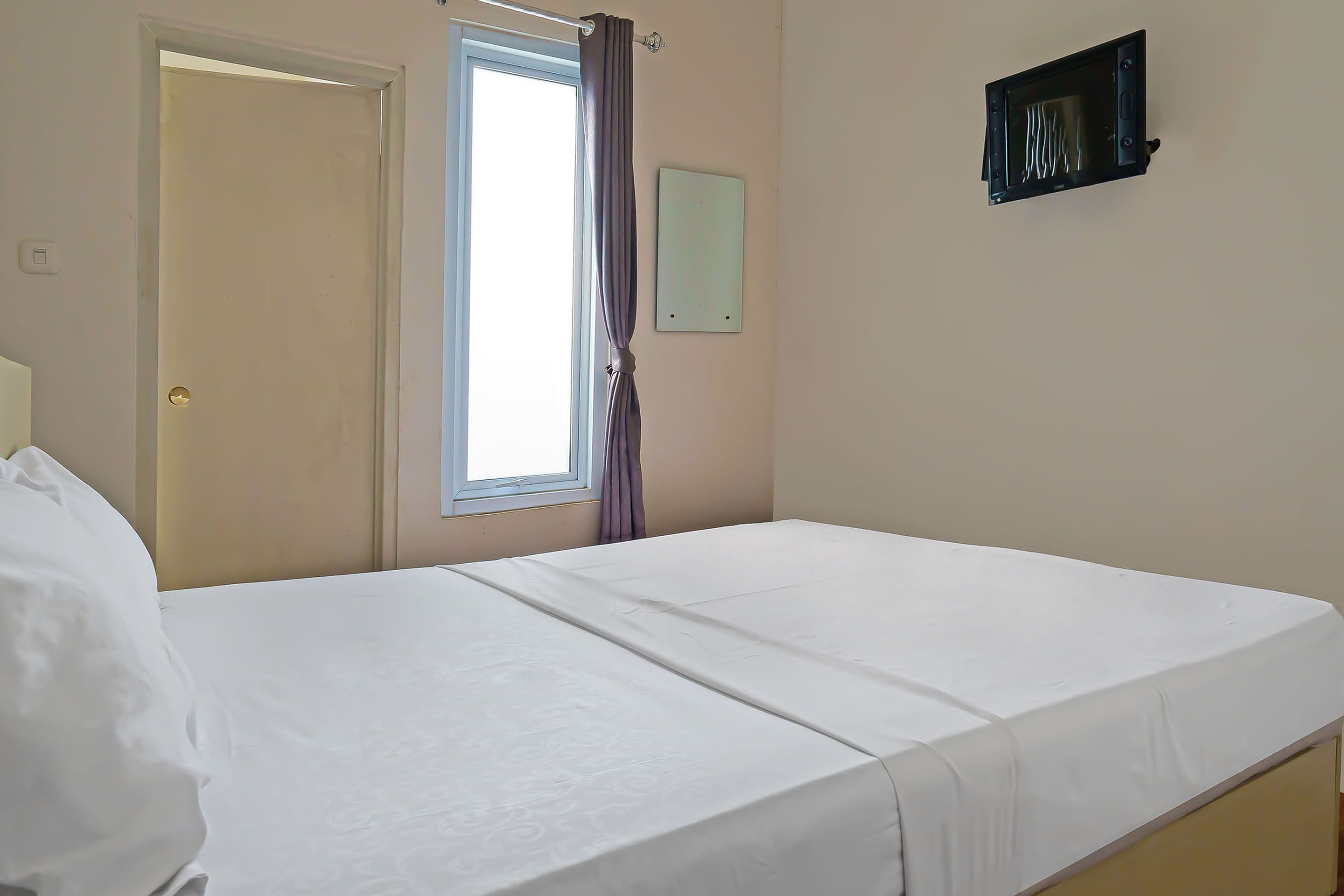 Ein schlichter, sauberer Raum mit einem Bett, einem Fenster mit Vorhängen und einem an der Wand montierten Fernseher.