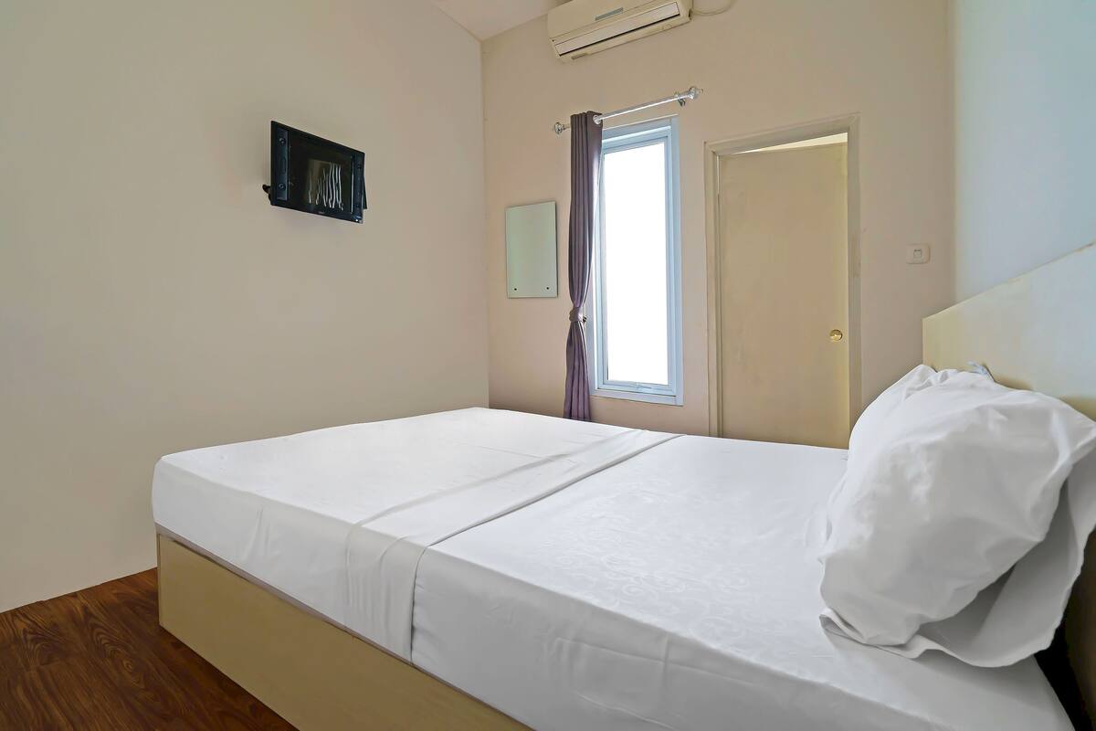 Un lit simple avec un matelas et une tête de lit blancs, une fenêtre avec des rideaux, une porte et un climatiseur fixé au mur.