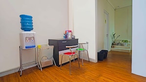 A room with a water dispenser, a desk with a chair, and a small table with a chair. There are potted plants and a trash bin in the corner. - OYO Life 92720 Mekarwangi Guest House (Bandung)