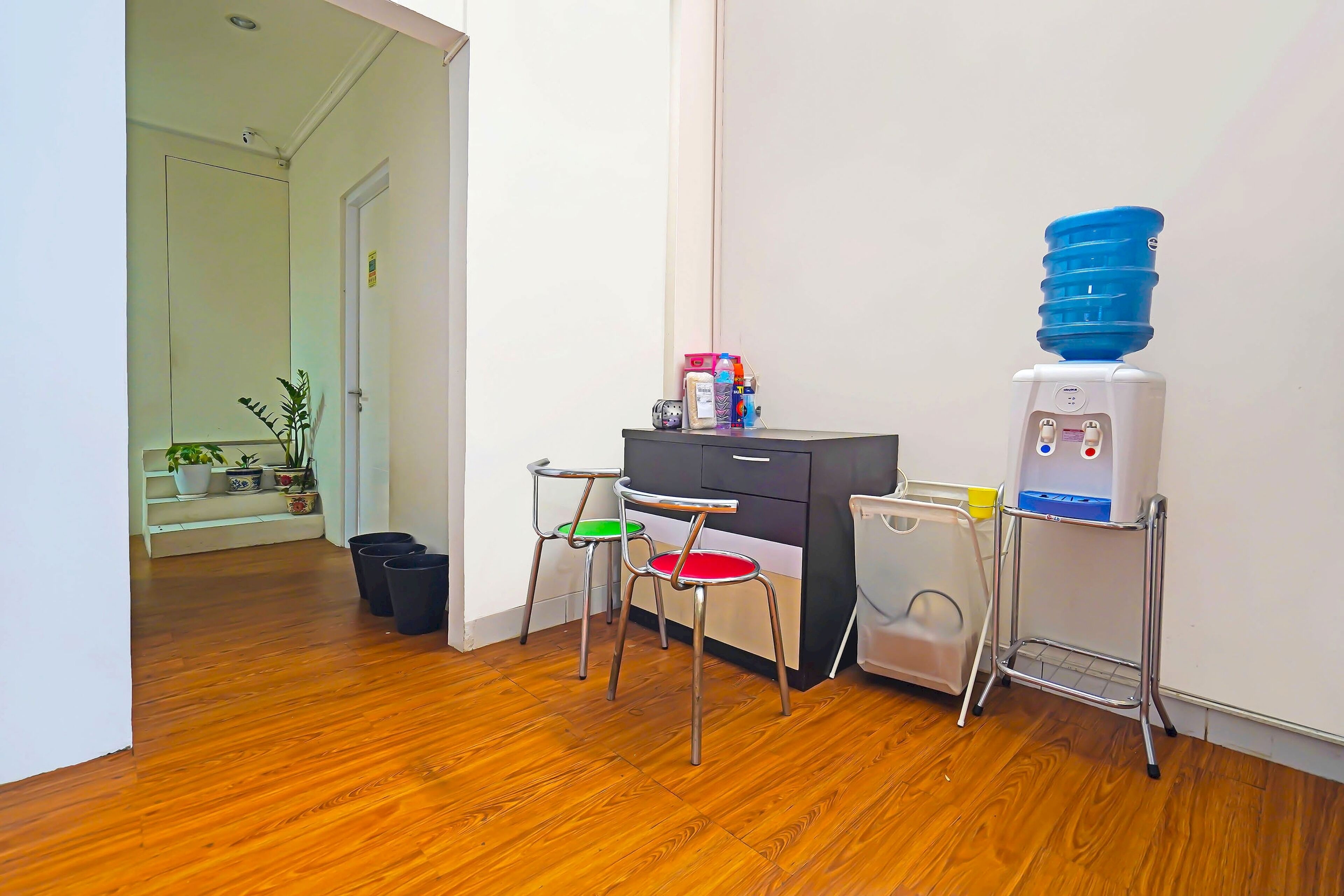 A room with wooden flooring, a desk with drawers, two chairs, a water dispenser, and a trash bin.