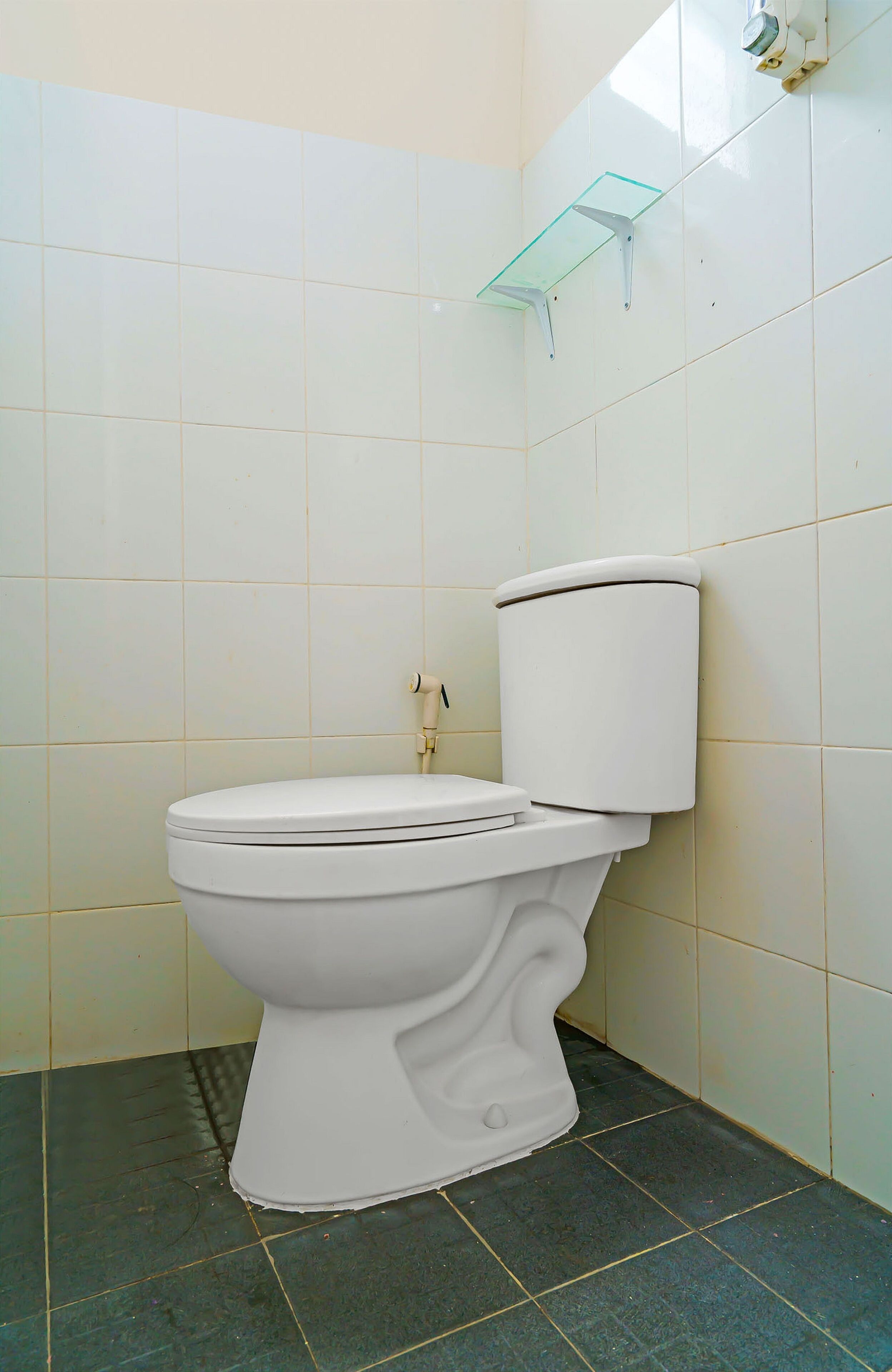 Des toilettes blanches, avec un réservoir et un abattant, une étagère en verre fixée au mur et un sol carrelé.