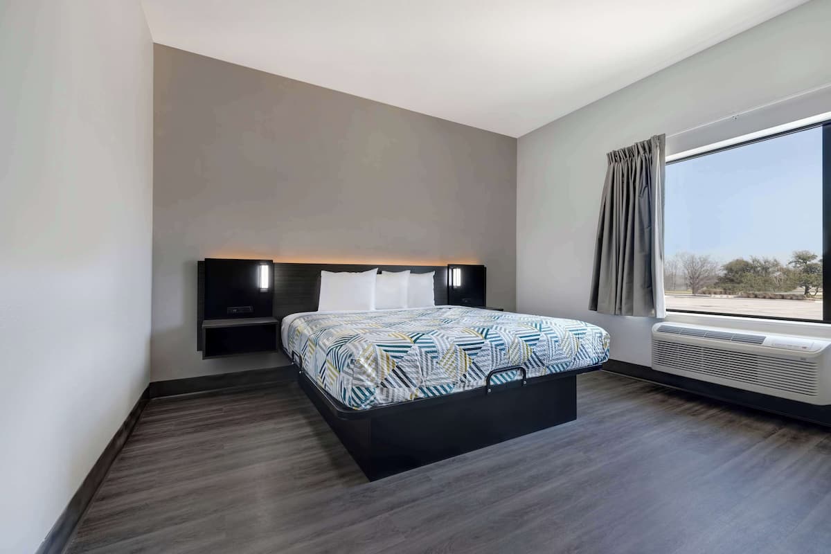 A modern hotel room with a large bed, a built-in TV, a bedside table, and a large window with curtains.