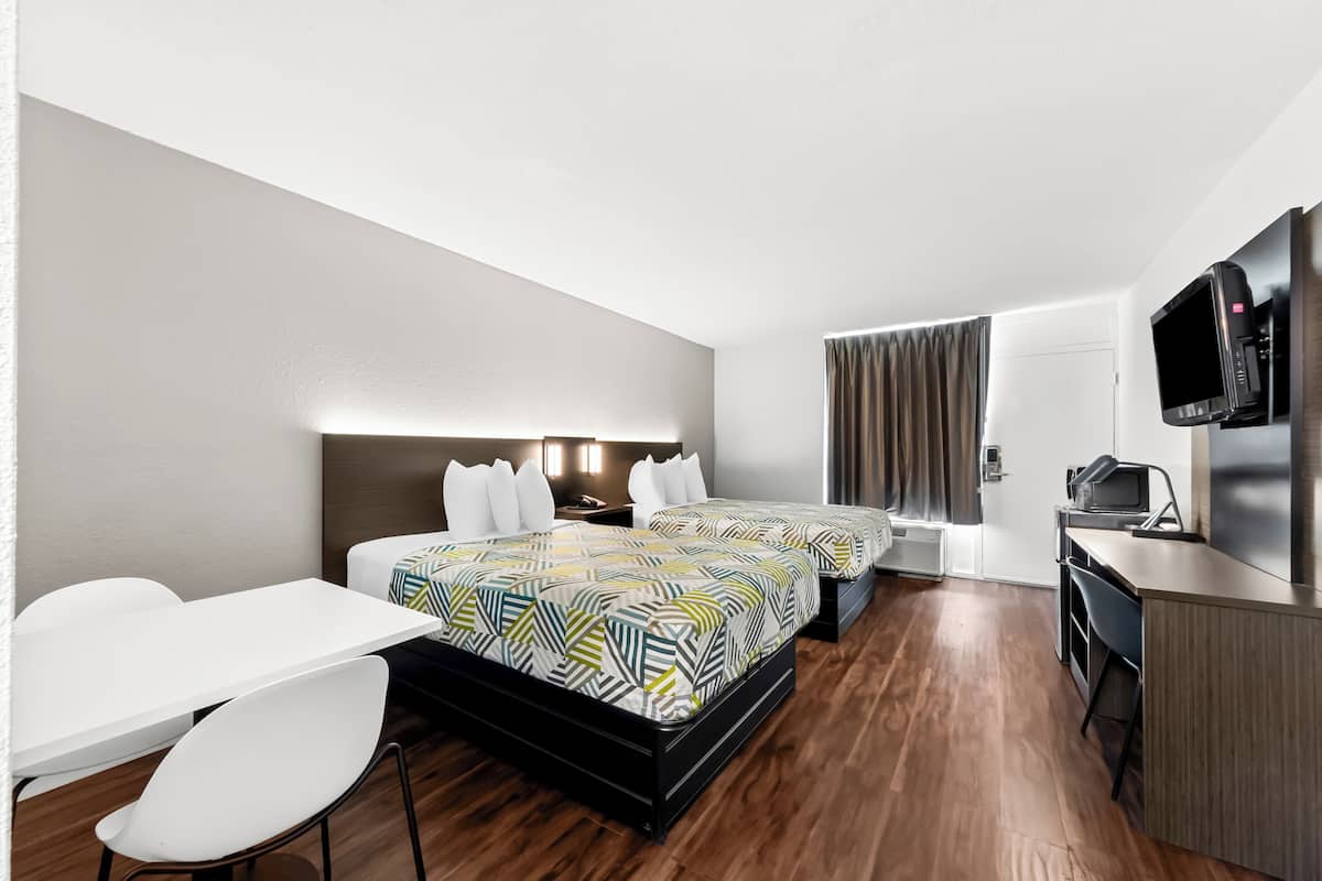 A hotel room with two beds, a desk, a chair, a TV, and a kitchenette.
