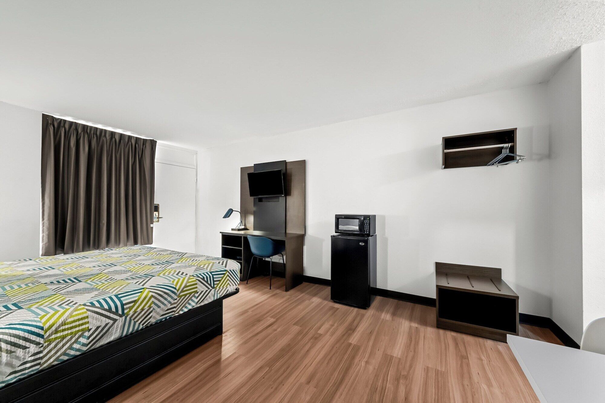 A hotel room with a bed, desk, chair, TV, microwave, and a small shelf.