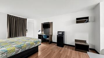 A hotel room with a bed, desk, chair, TV, microwave, and a small shelf.
