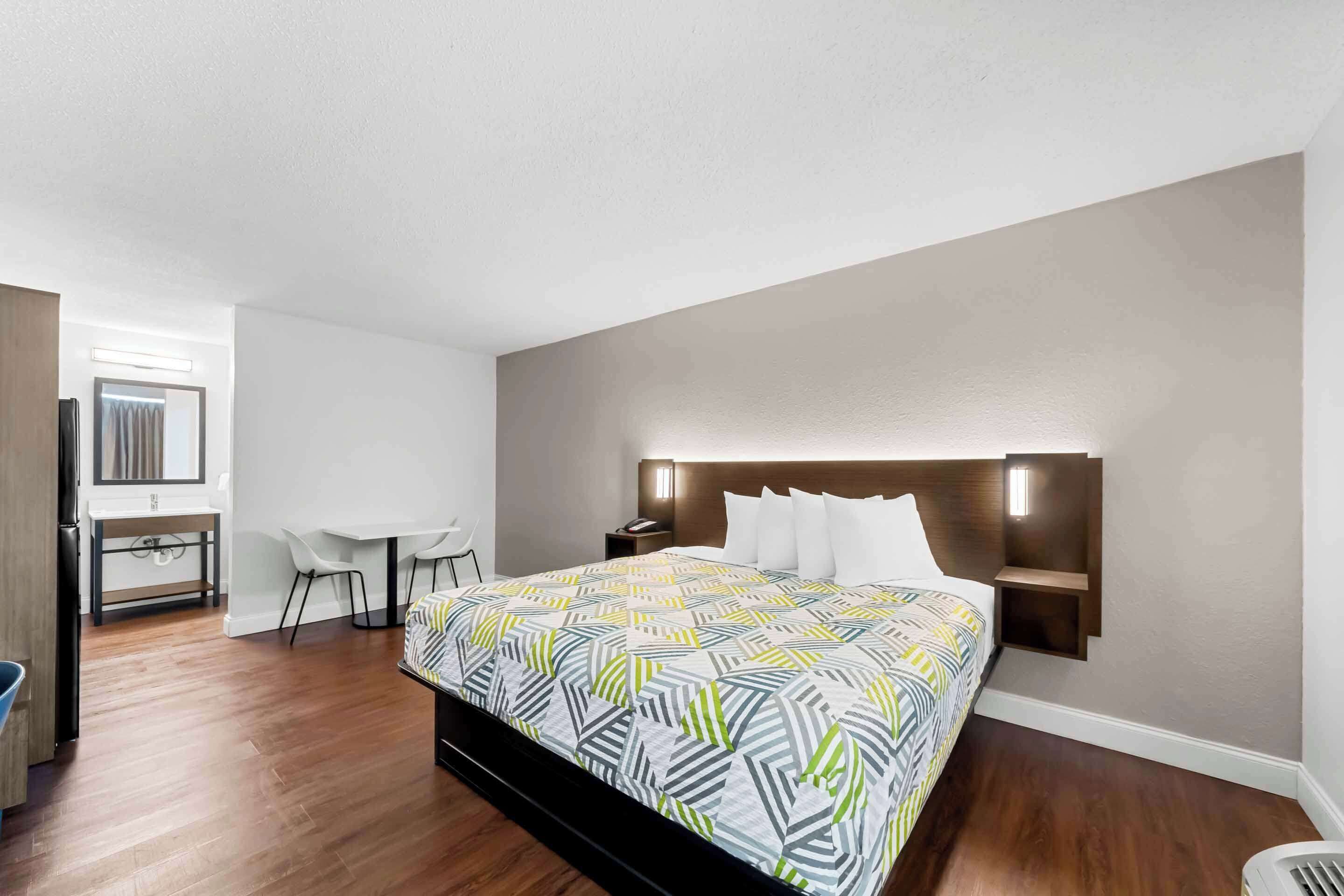 A hotel room with a large bed, a desk with a chair, a small table, and a wall-mounted nightstand.