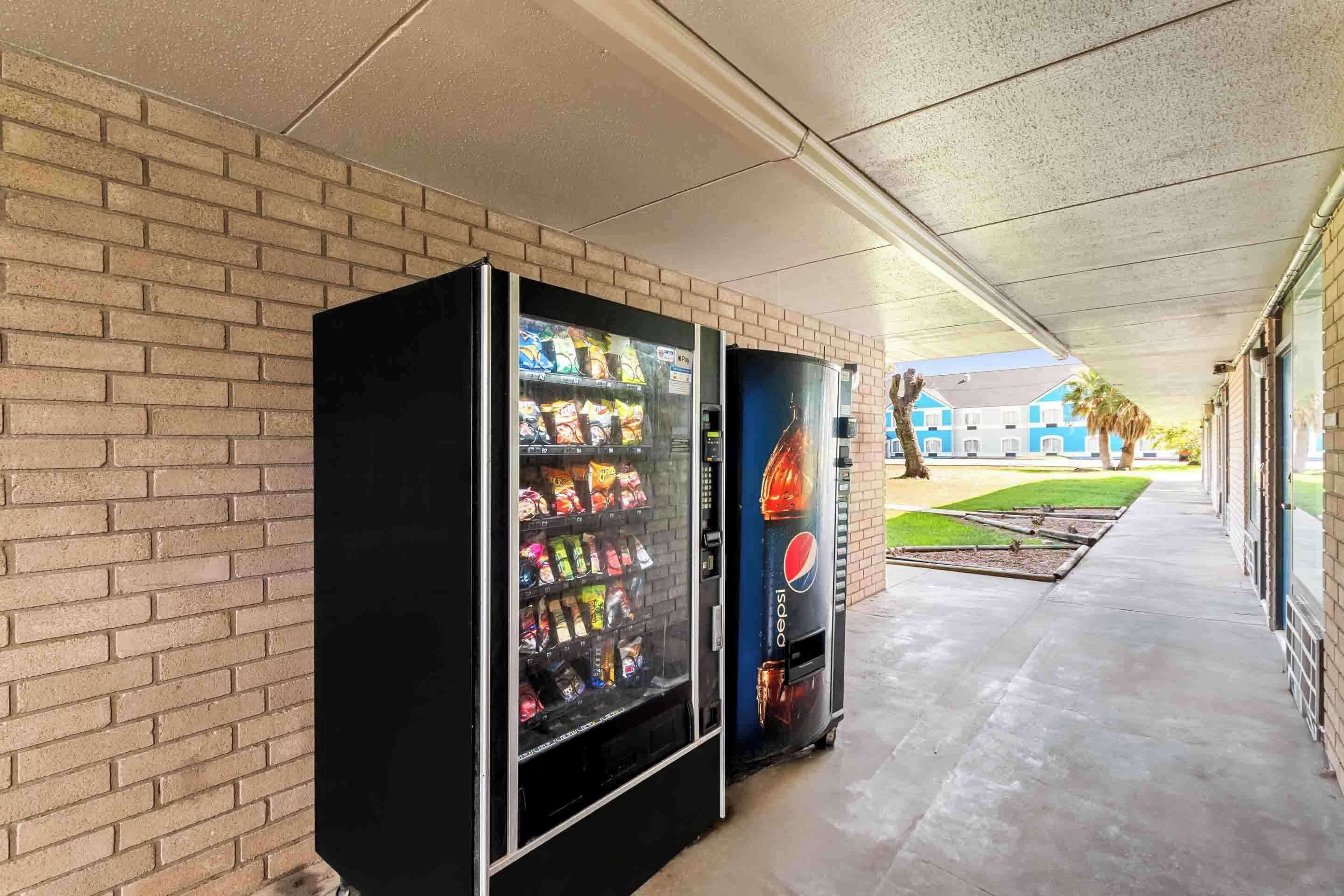 A vending machine and a Pepsi refrigerator in an outdoor area with a brick wall and a covered walkway.