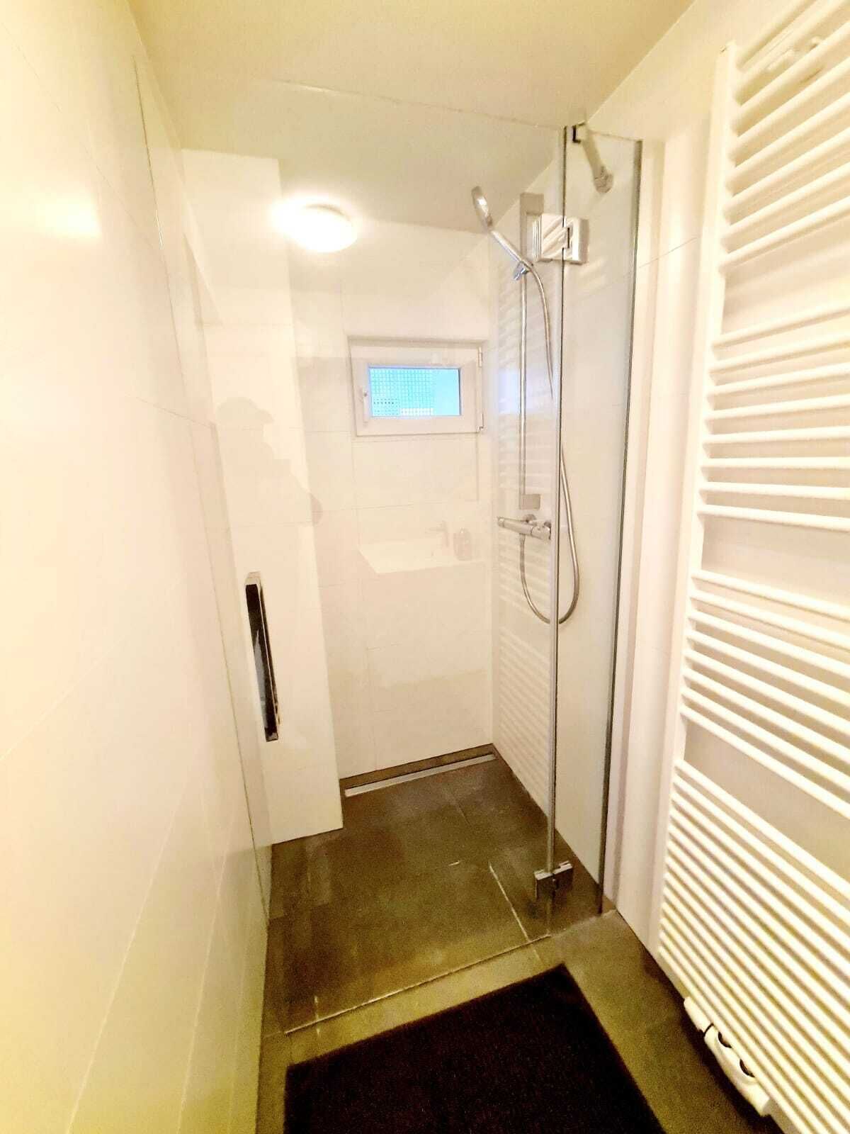 A small, enclosed shower area with a glass door, a window, and a radiator on the right.