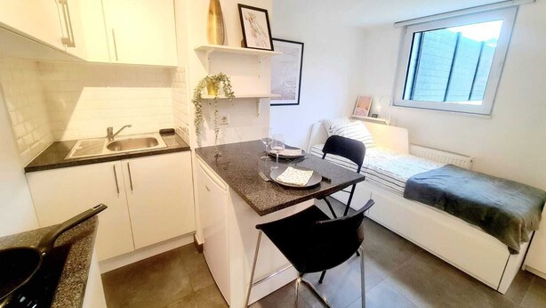 A compact kitchen with a sink, a dining area with a chair, a bed, and a window. - New Studio in Center (Luxembourg City)