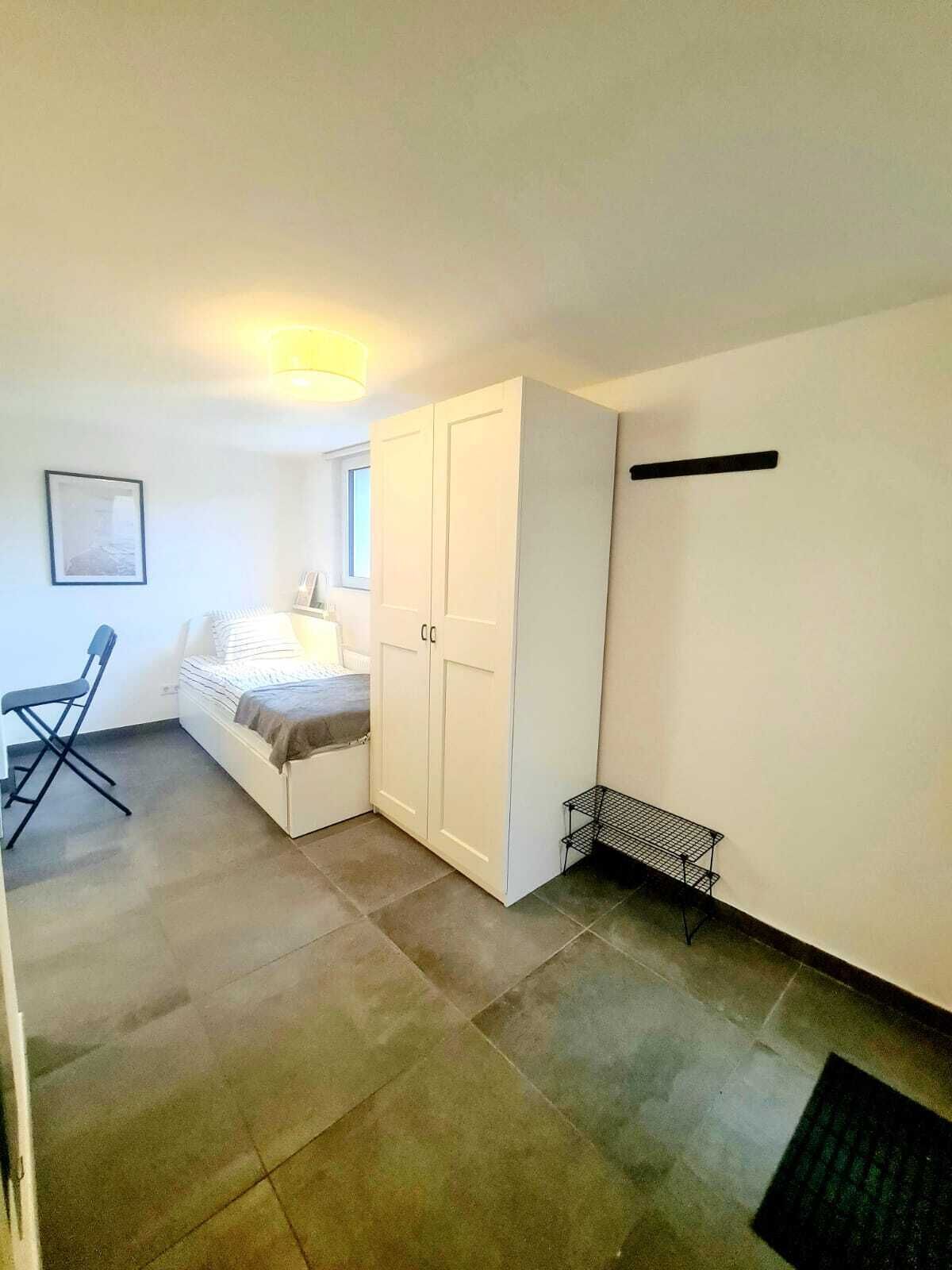 A small, well-lit room with a bed, a desk, and a wardrobe.