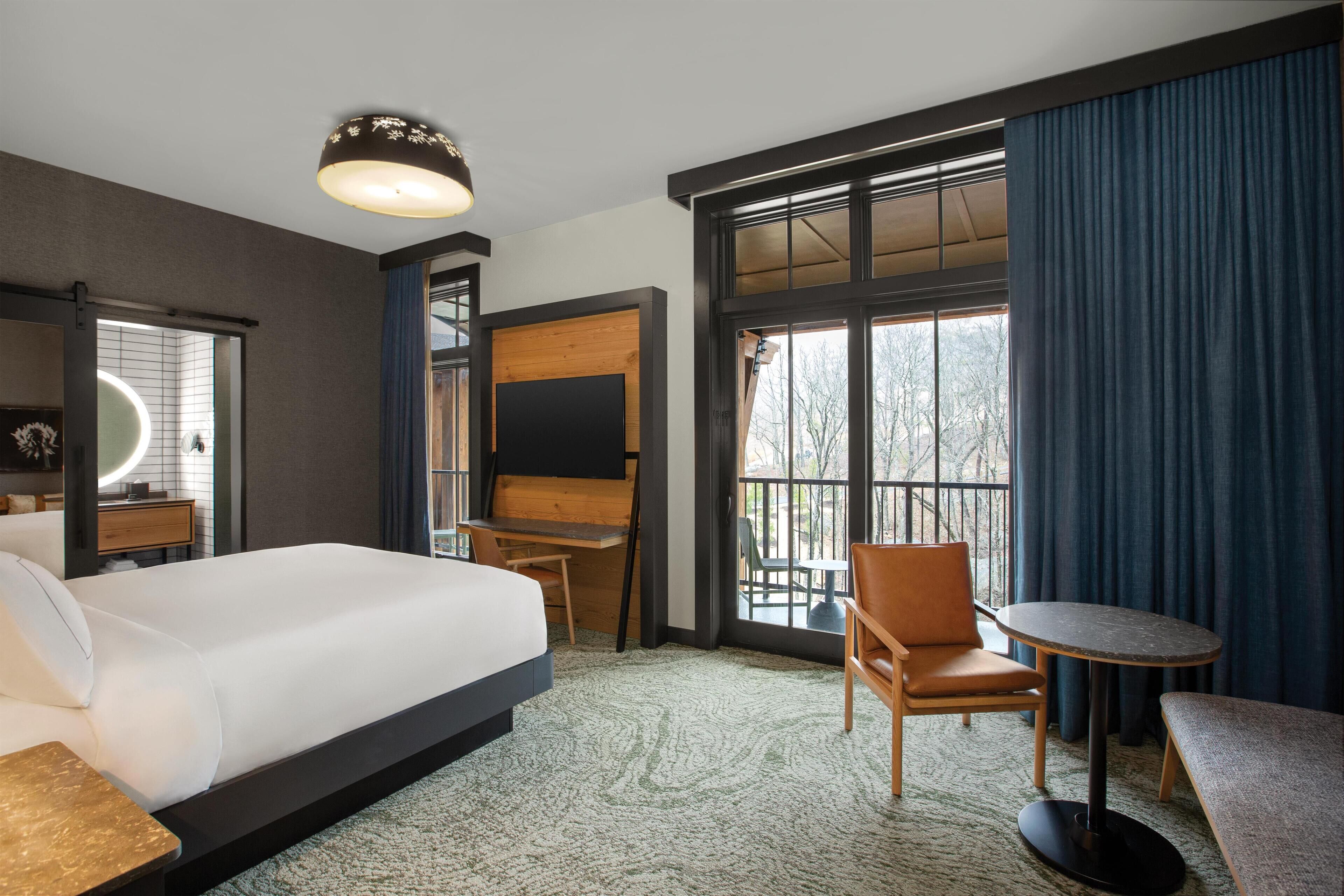 A hotel room with a large bed, a desk, a chair, a small table, and a large window with curtains.