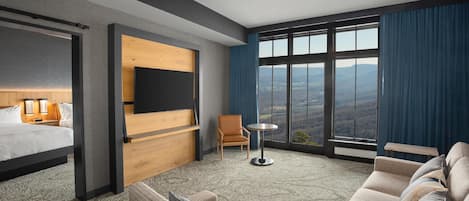 A hotel room with a bed, a sofa, a TV mounted on the wall, a small table, and a view of the mountains through large windows.