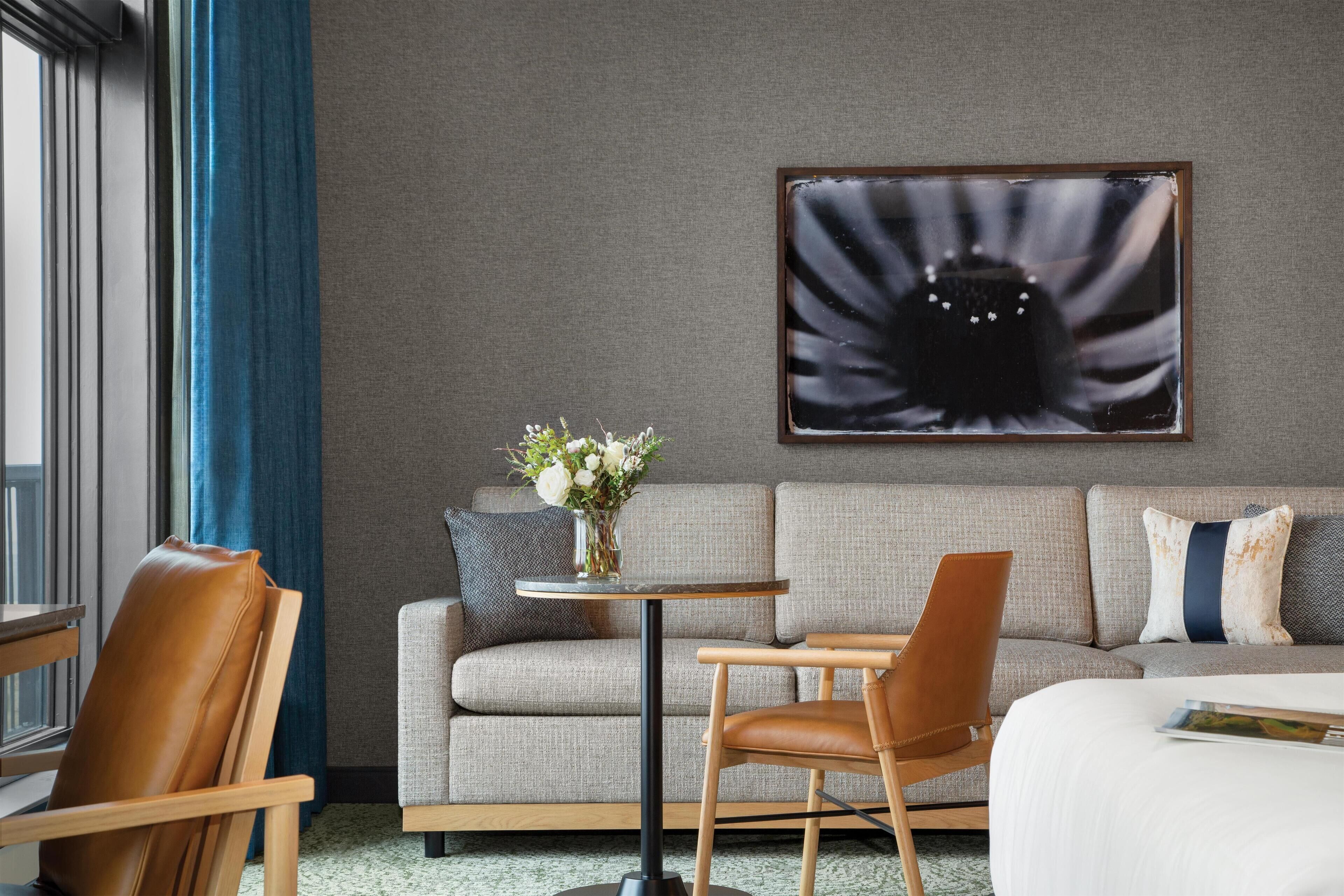 Hotellihuone, jossa on harmaa sohva, pieni pyöreä pöytä, jossa on maljakko täynnä kukkia, ruskea nahkainen nojatuoli ja valkoisella peitteellä varustettu sänky.