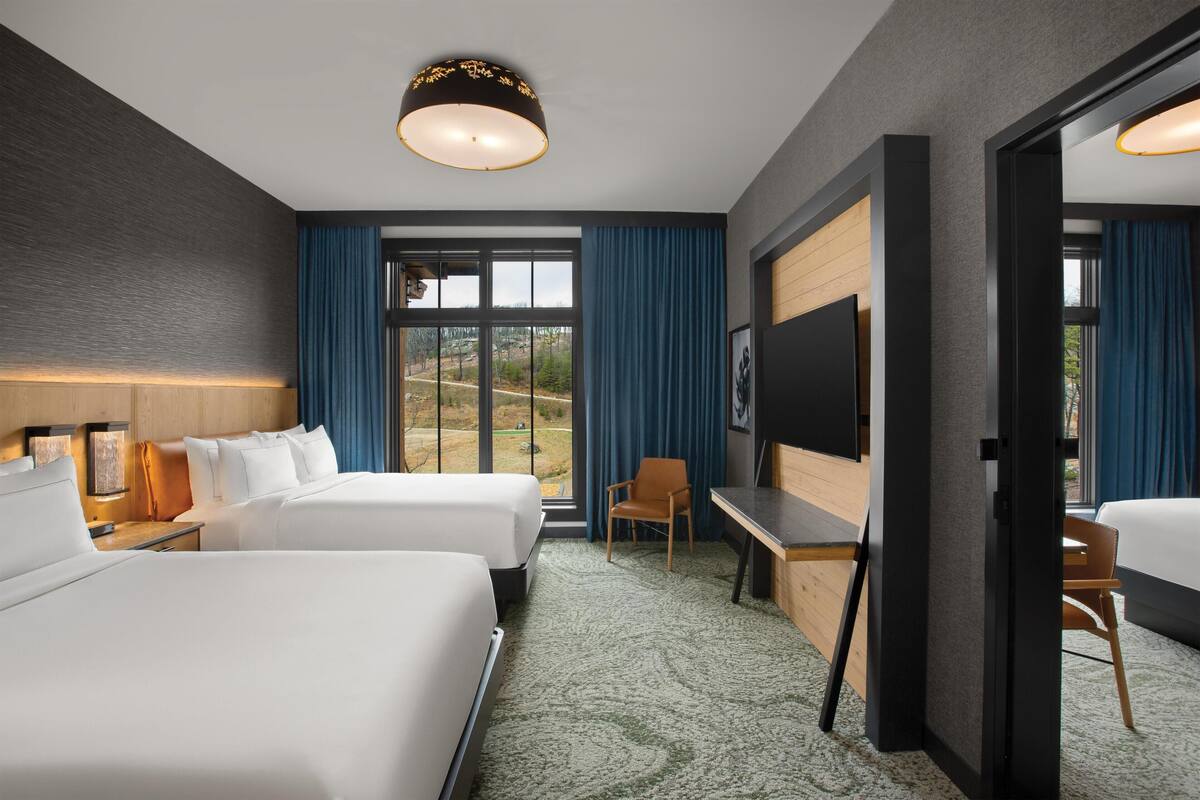A hotel room with two beds, a desk, a chair, a TV, and a large window with curtains.