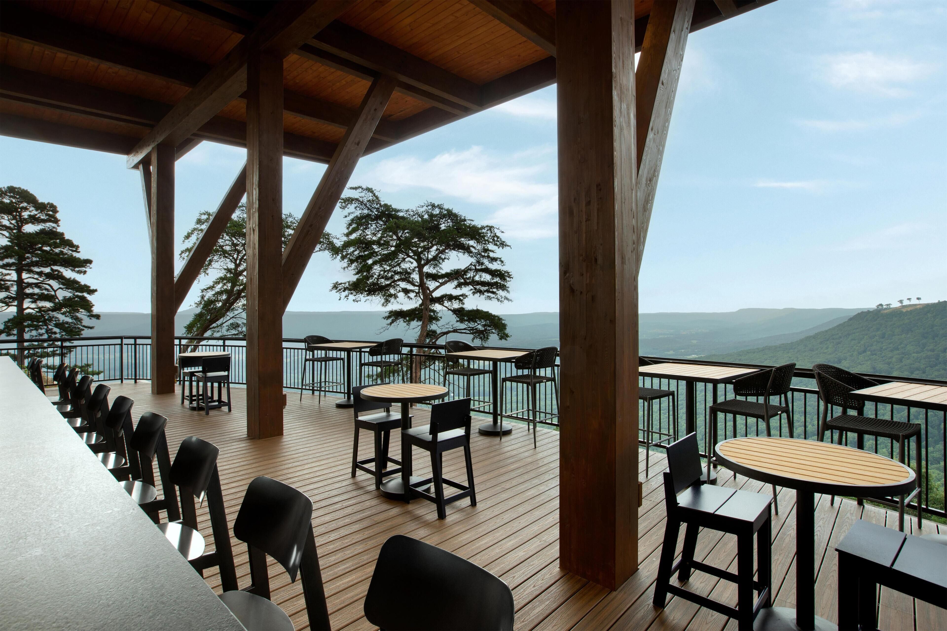 Una terraza al aire libre con mesas y sillas de madera, con vistas a un paisaje pintoresco con árboles y colinas.