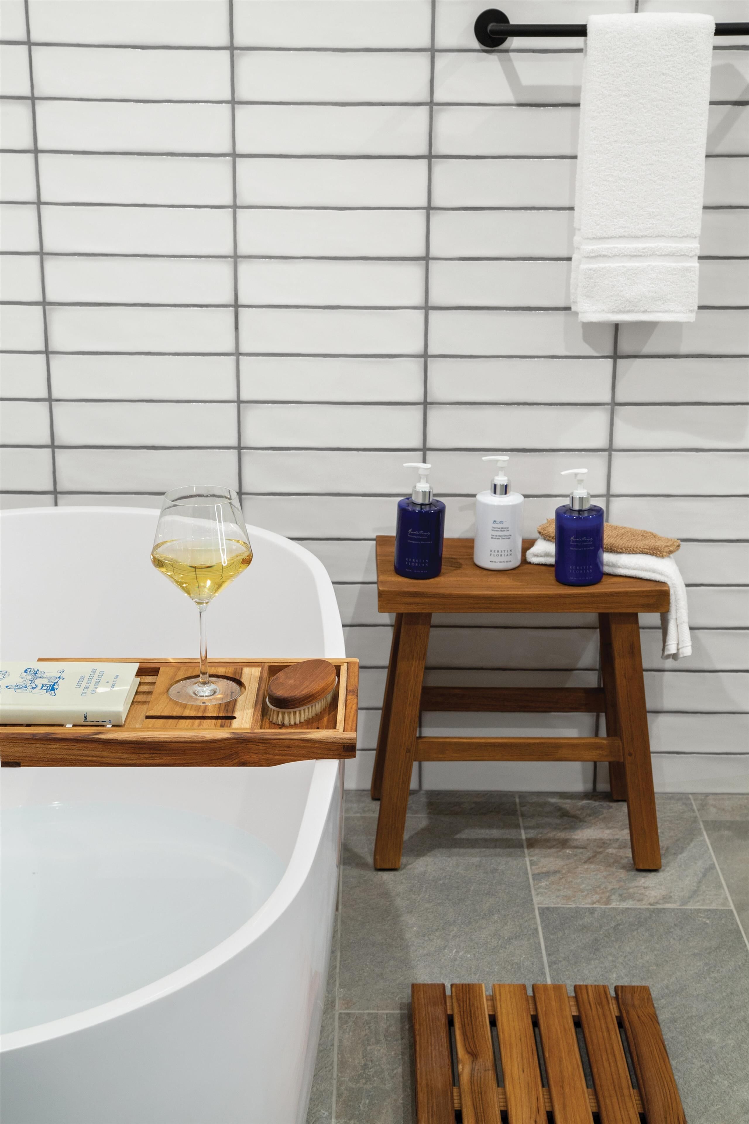 A modern bathroom with a freestanding bathtub, a wooden tray with a glass of wine, and toiletries on a small wooden table.