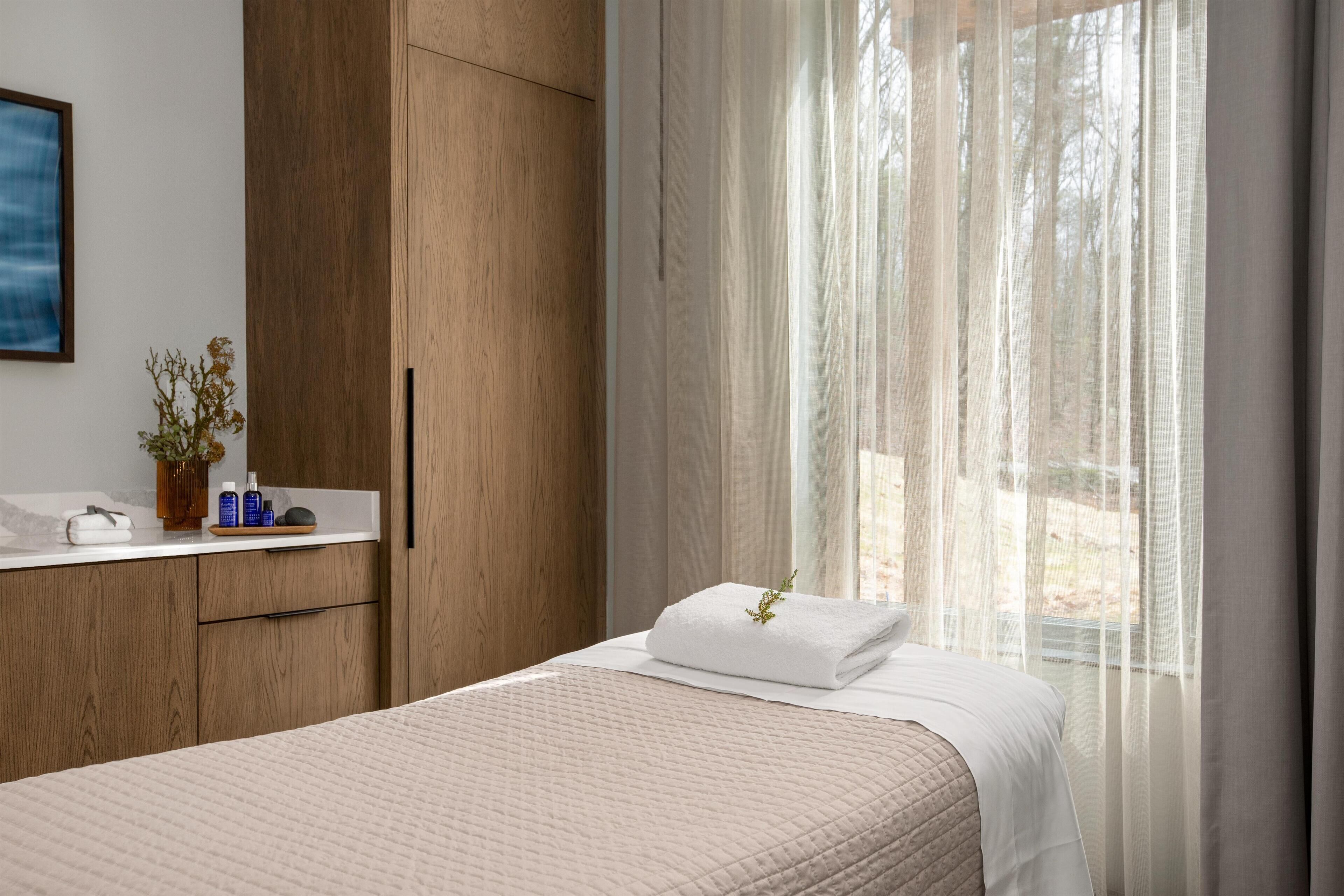 Un lit bien fait, avec un oreiller blanc et une serviette blanche pliée, une armoire en bois, une petite table avec des articles de toilette et une fenêtre ornée de rideaux légers.
