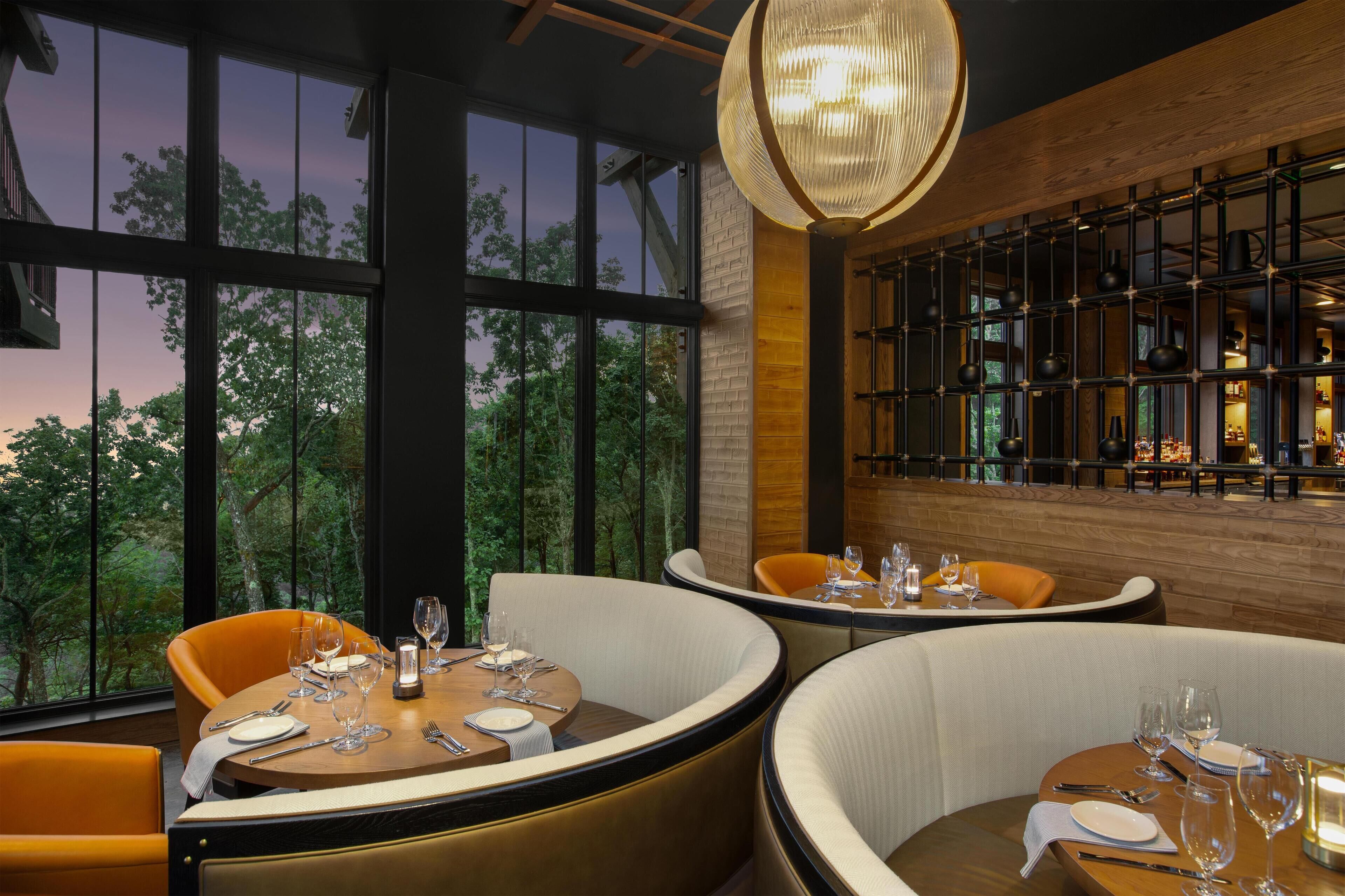 Un ristorante con tavoli rotondi e sedie imbottite, ampie finestre che offrono vista su alberi, e un design moderno con mattoni a vista e travi in legno.