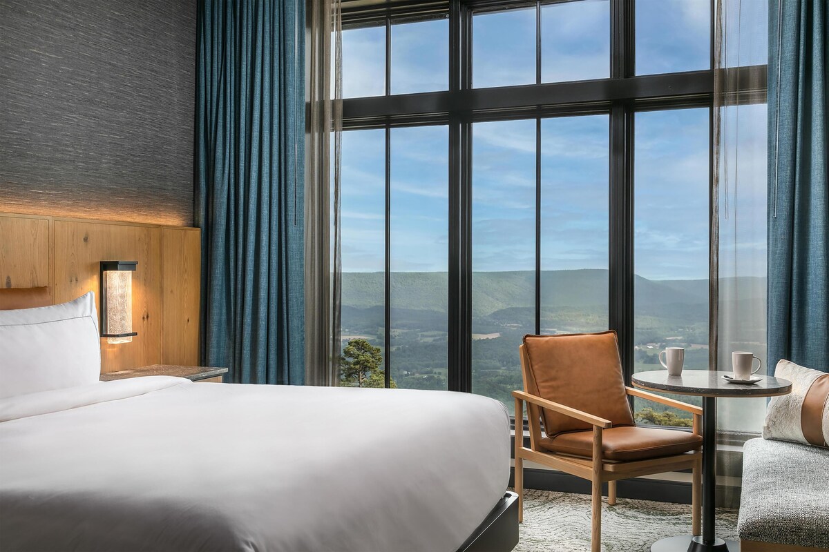 Habitación de hotel con un ventanal grande, una cama, una silla y una mesita con tazas.