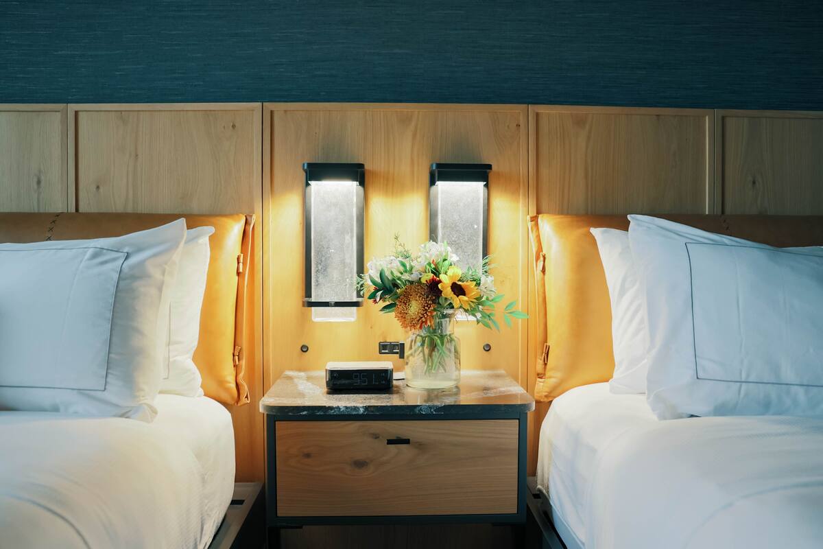A hotel room with two beds, a nightstand with a clock and a vase of flowers, and wooden paneling on the walls.