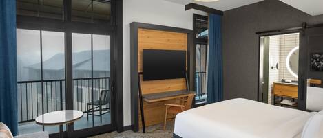 A hotel room with a large bed, a desk with a chair, a TV, a small table, and a balcony with a view of the mountains.