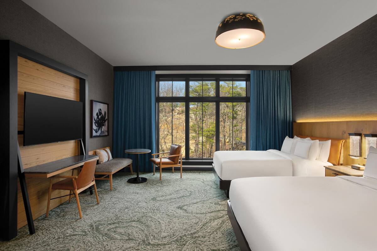 A hotel room with two beds, a desk, a chair, a TV, and a large window with curtains.