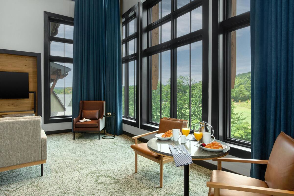 A hotel room with a large window, a dining table with breakfast, a TV mounted on the wall, and a cozy seating area.