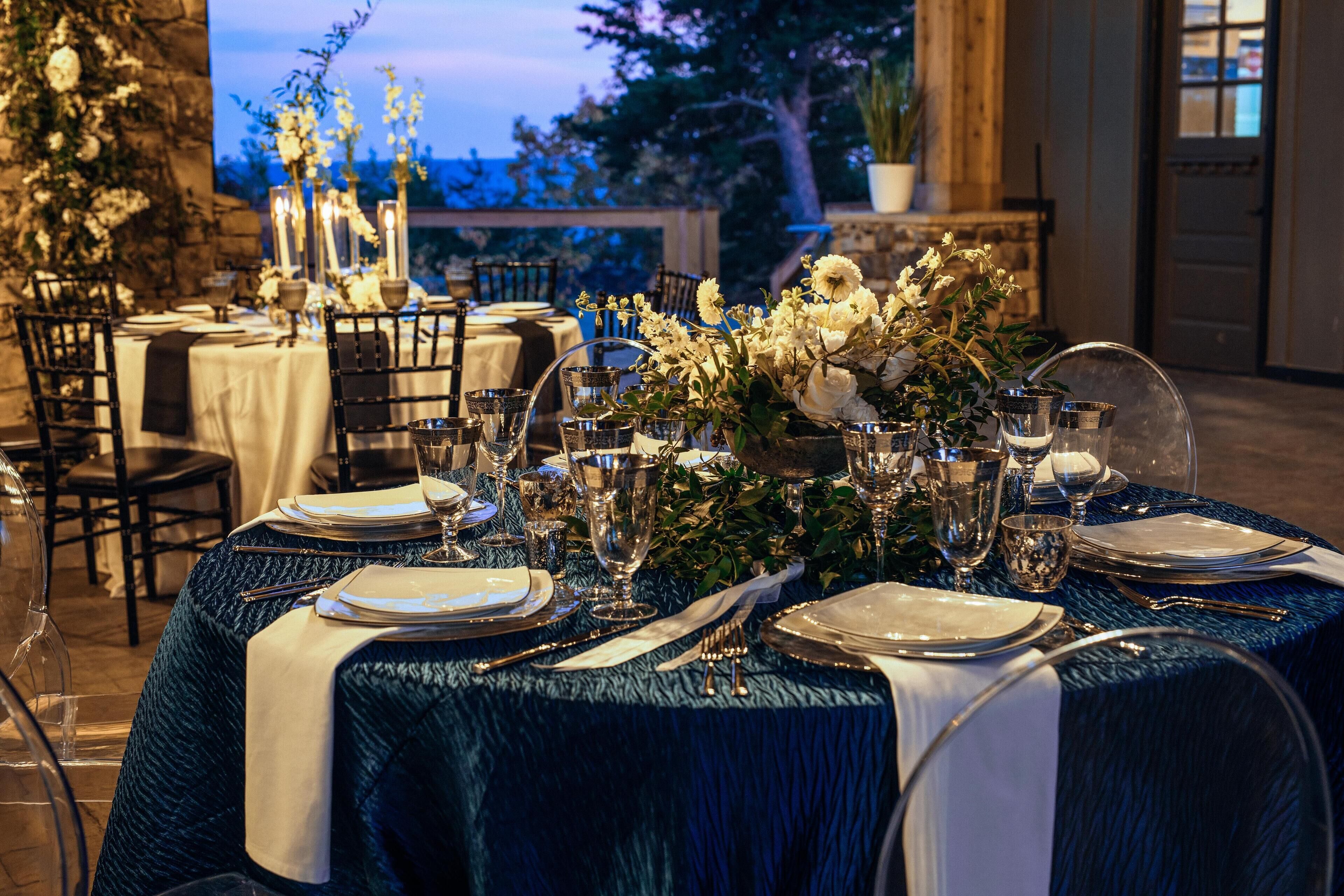 A table set for a formal outdoor dining experience with white tablecloths, silverware, and floral centerpieces.