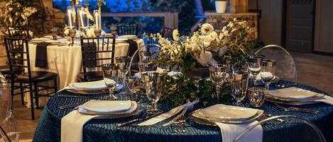 A table set for a formal outdoor dining experience with white tablecloths, silverware, and floral centerpieces.