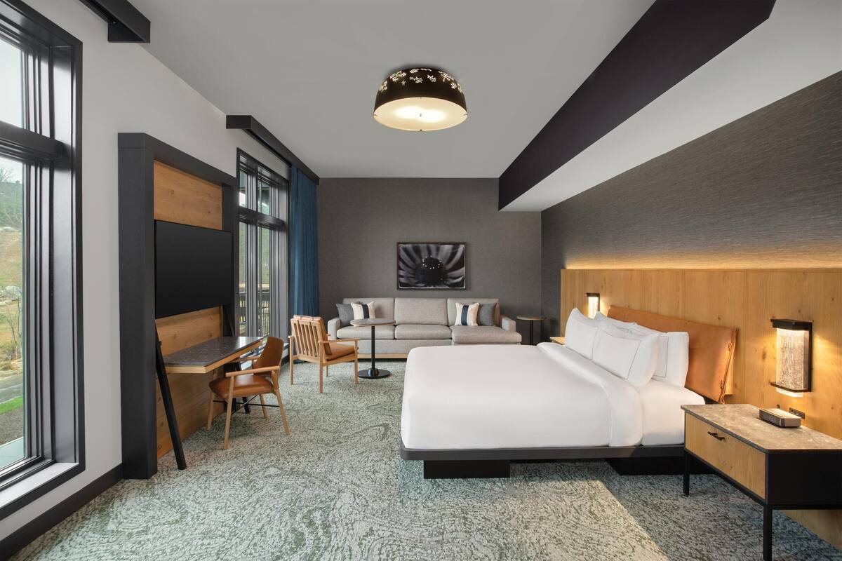 A modern hotel room with a large bed, a desk, a sofa, and a view of the outdoors.