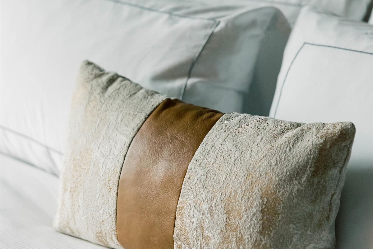 A bed with white linens and a single decorative pillow with a leather accent.