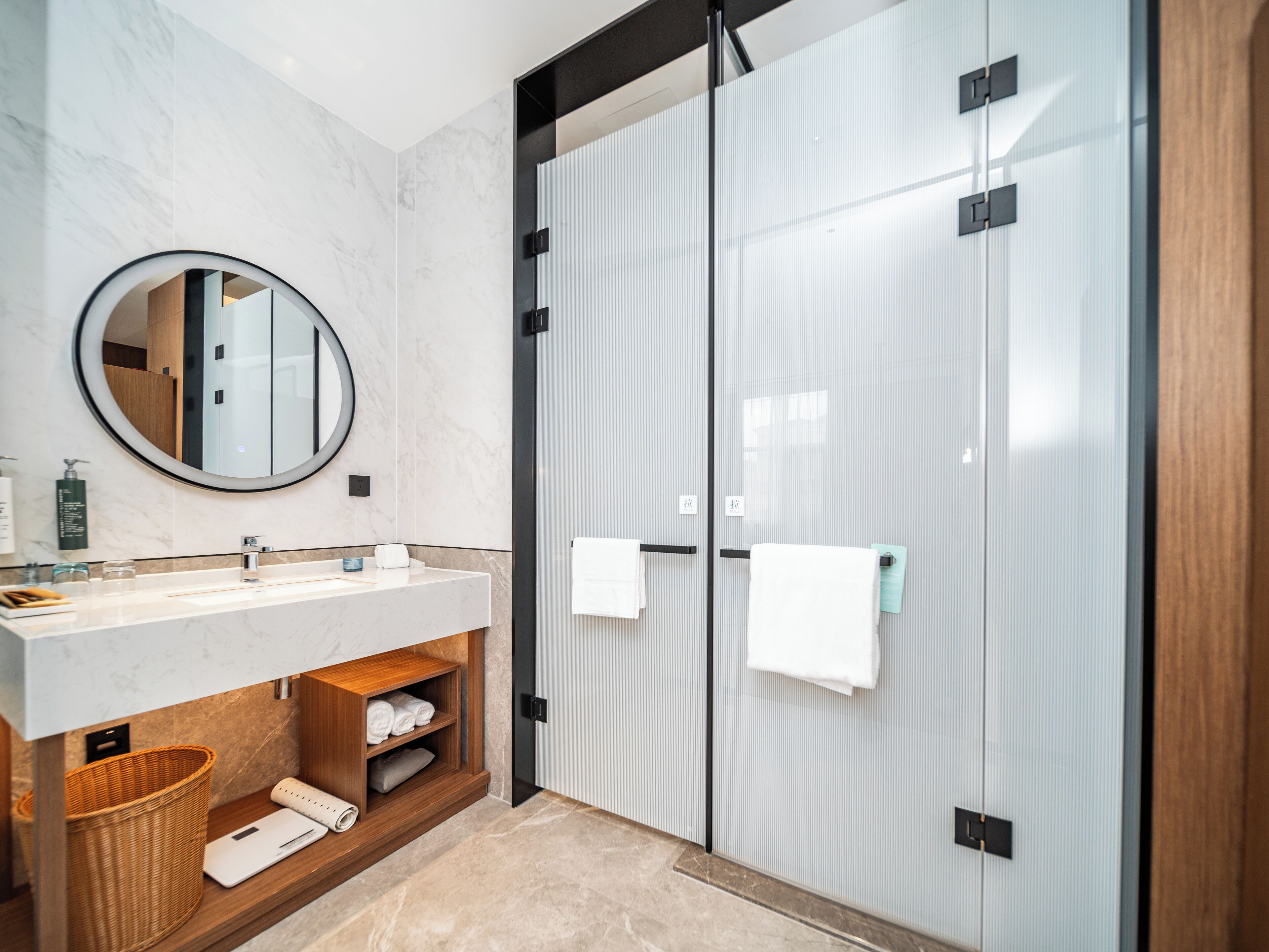 Un baño moderno con un lavamanos de mármol, un espejo redondo, cabina de ducha de vidrio y un estante de madera con toallas.