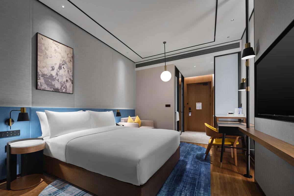Una habitación de hotel moderna con una cama grande, mesitas de noche, un escritorio con una silla, un espejo amplio y un cuadro en la pared.