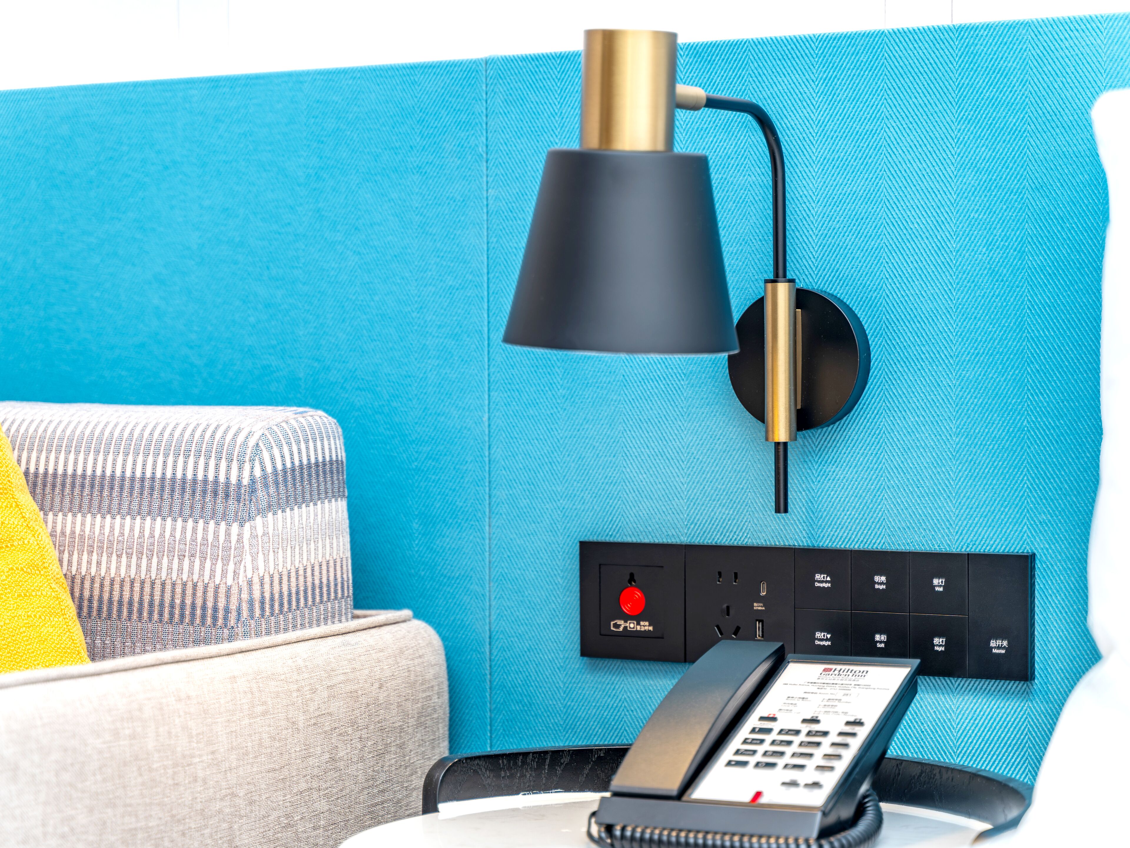 Una habitación de hotel moderna con una pared azul con textura, una lámpara negra, un teléfono y una regleta con múltiples enchufes.