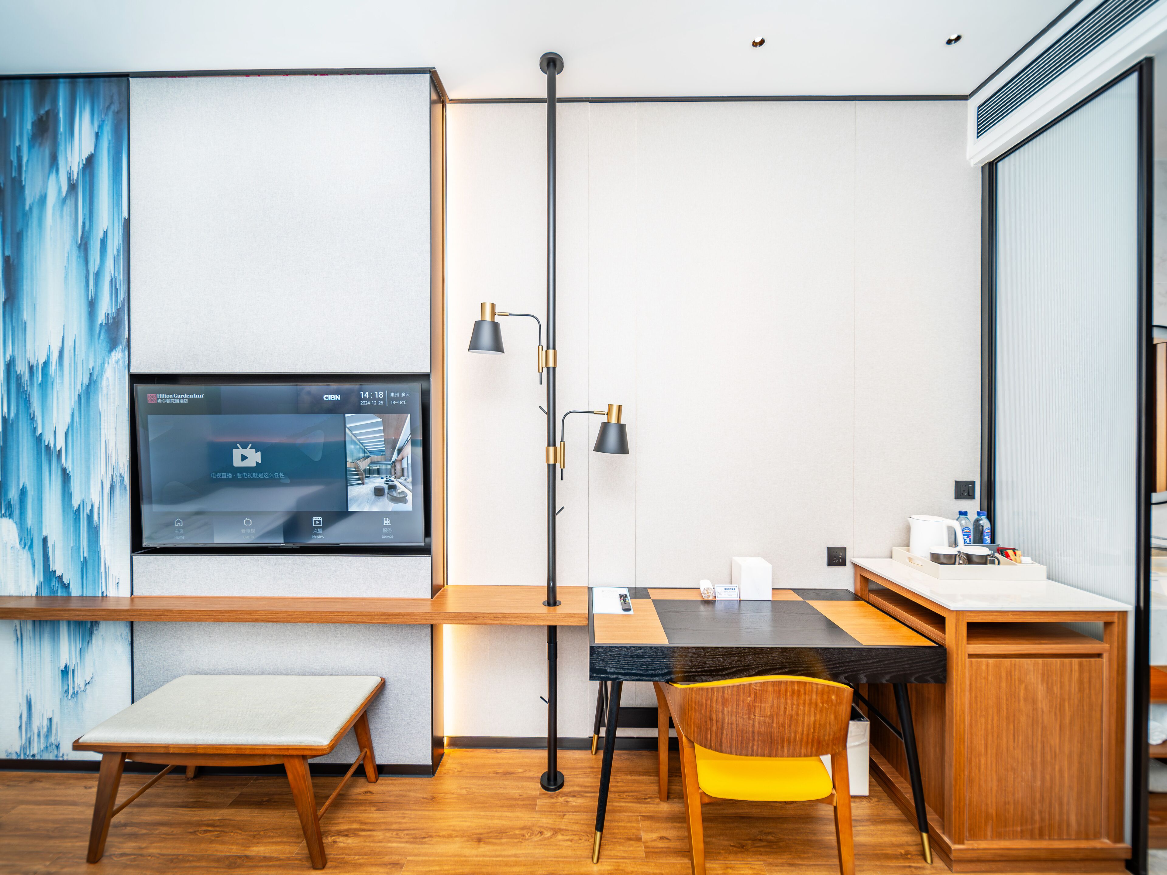 A modern hotel room with a wooden desk, a bench, a yellow chair, a flat-screen TV mounted on the wall, and a large window.