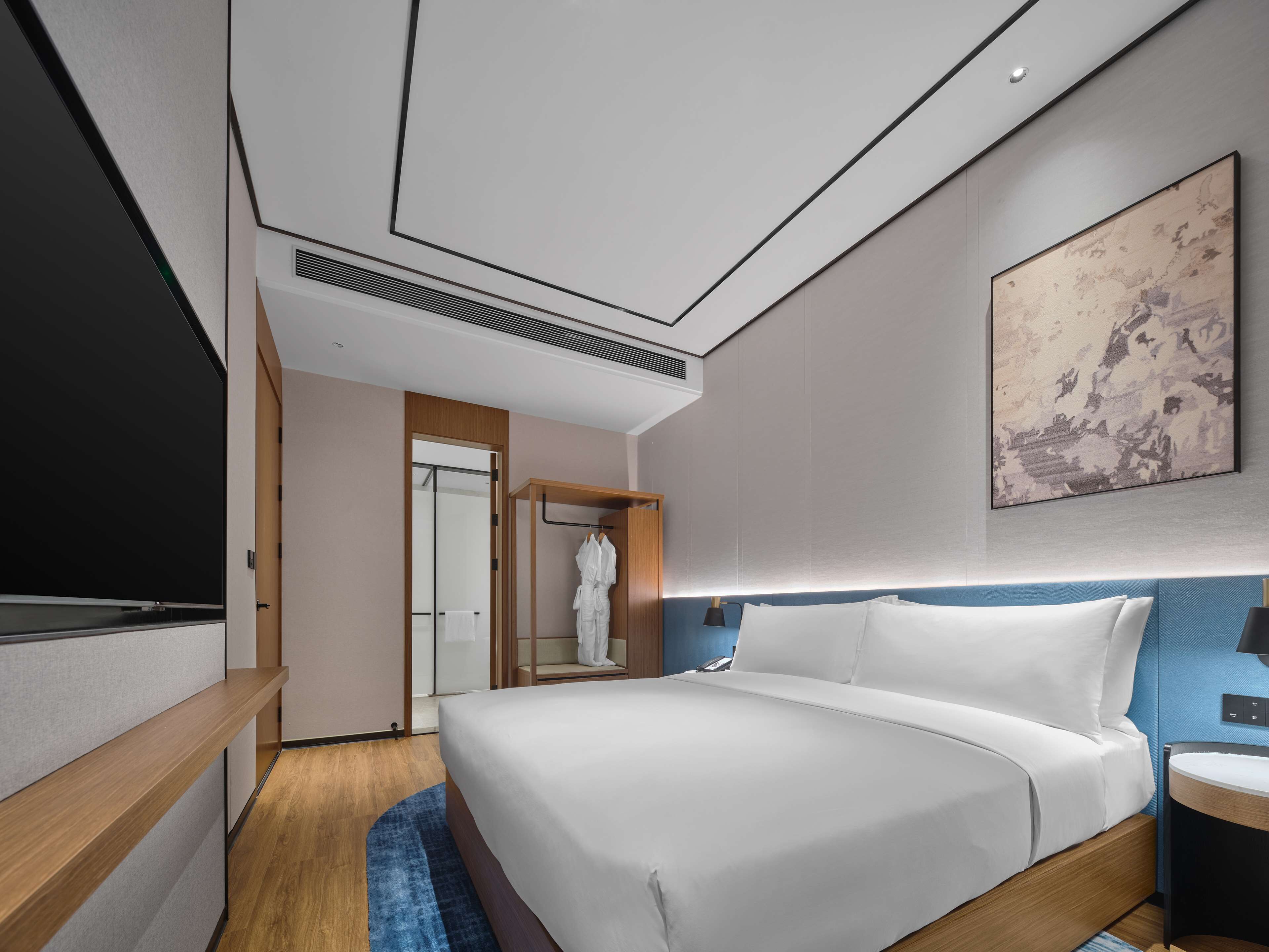 A modern hotel room with a large bed, a bedside table, a TV, and a bathroom visible through an open door.