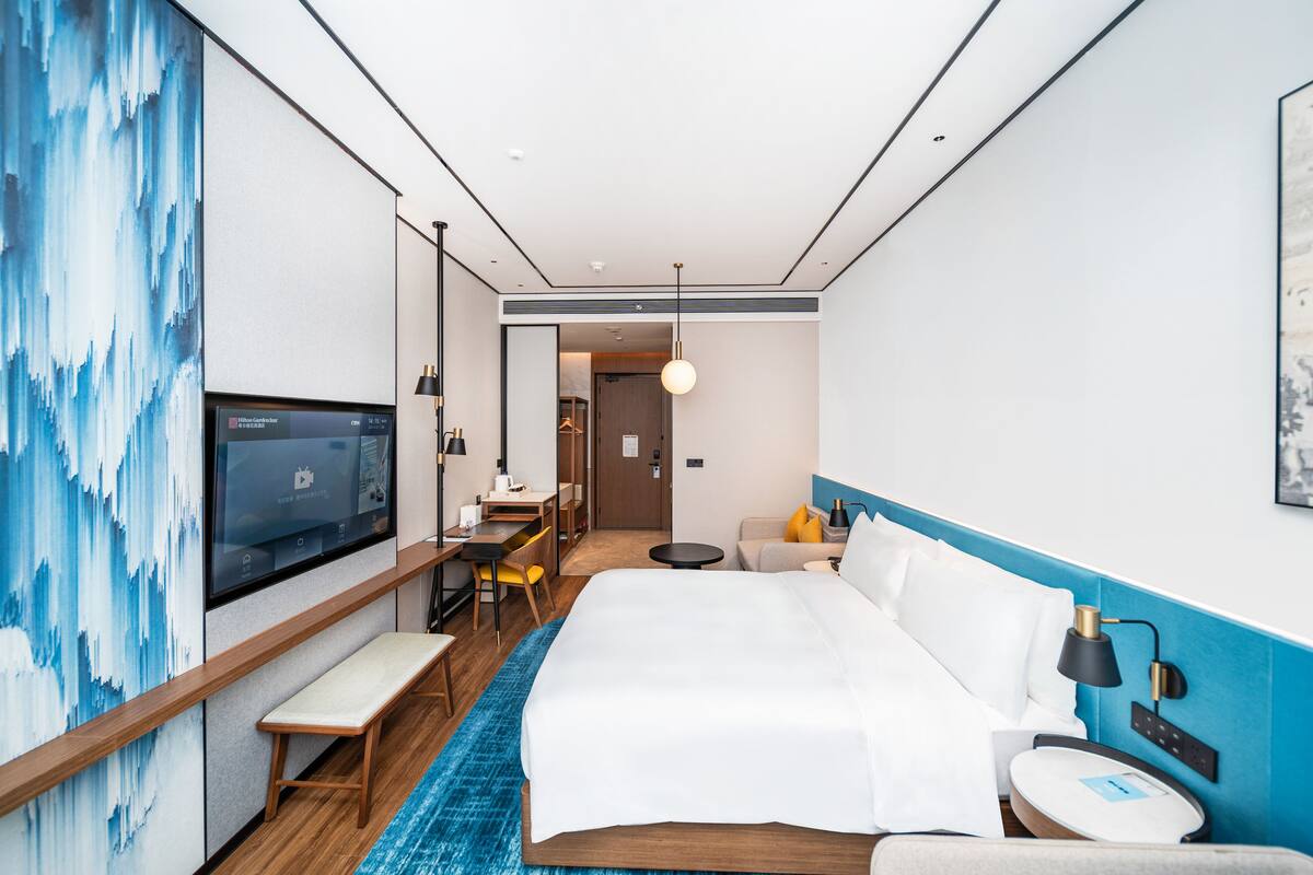 Una habitación de hotel moderna con una cama grande, un escritorio con silla, un televisor y un baño a la vista al fondo.