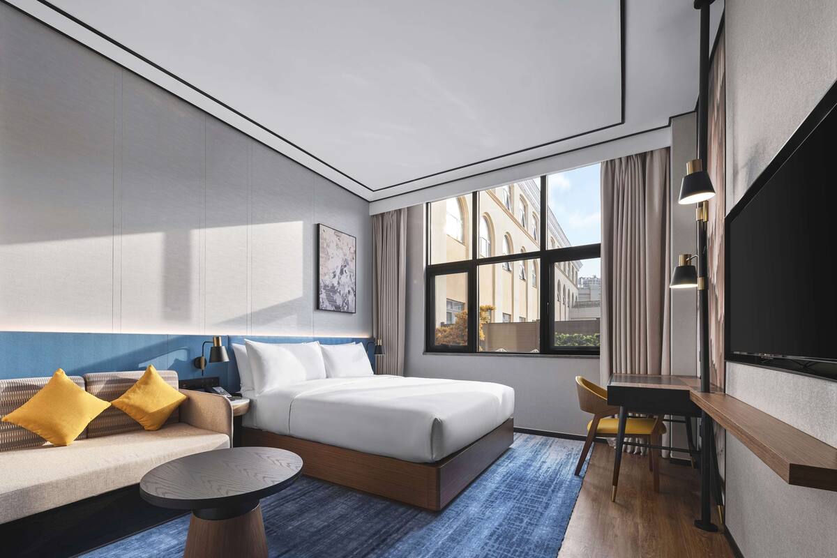 Una habitación de hotel moderna con una cama grande, un sofá con cojines amarillos, una mesita redonda pequeña y un escritorio con una silla.
