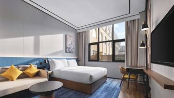 A modern hotel room with a large bed, a sofa with yellow pillows, a small round table, and a desk with a chair.