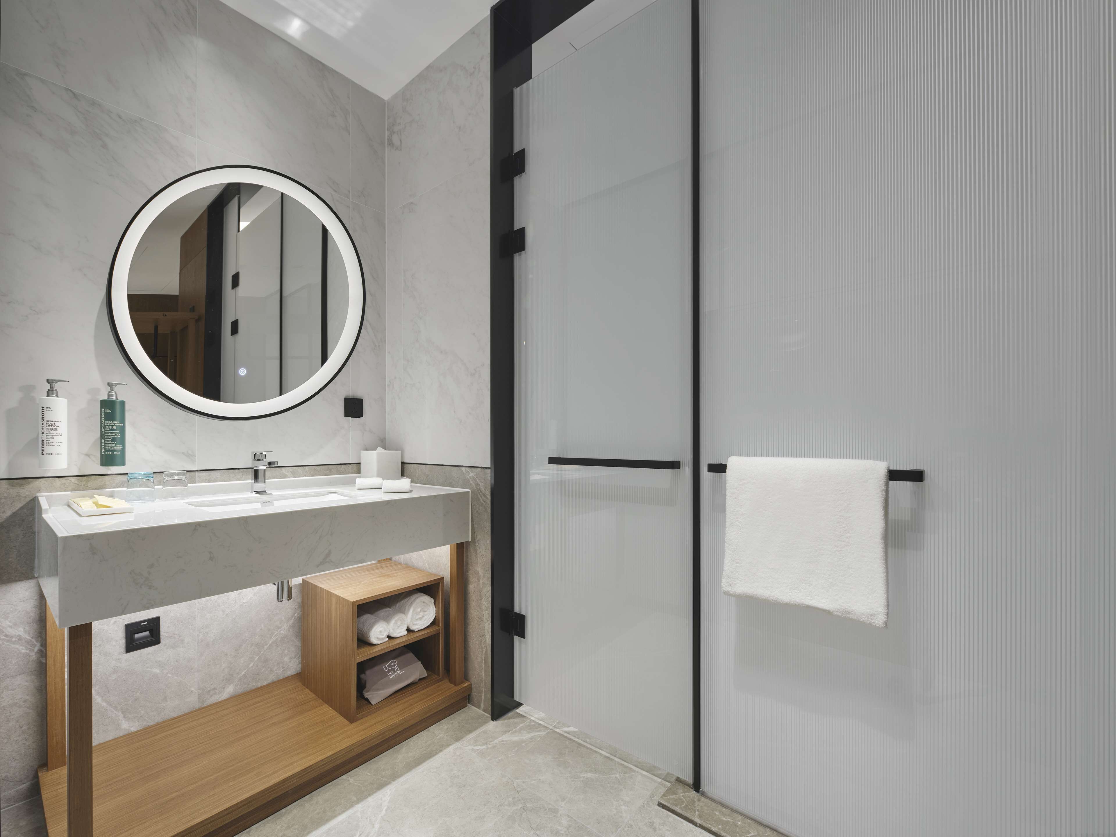 A modern bathroom with a marble sink, a round mirror, a glass shower enclosure, and a wooden vanity.
