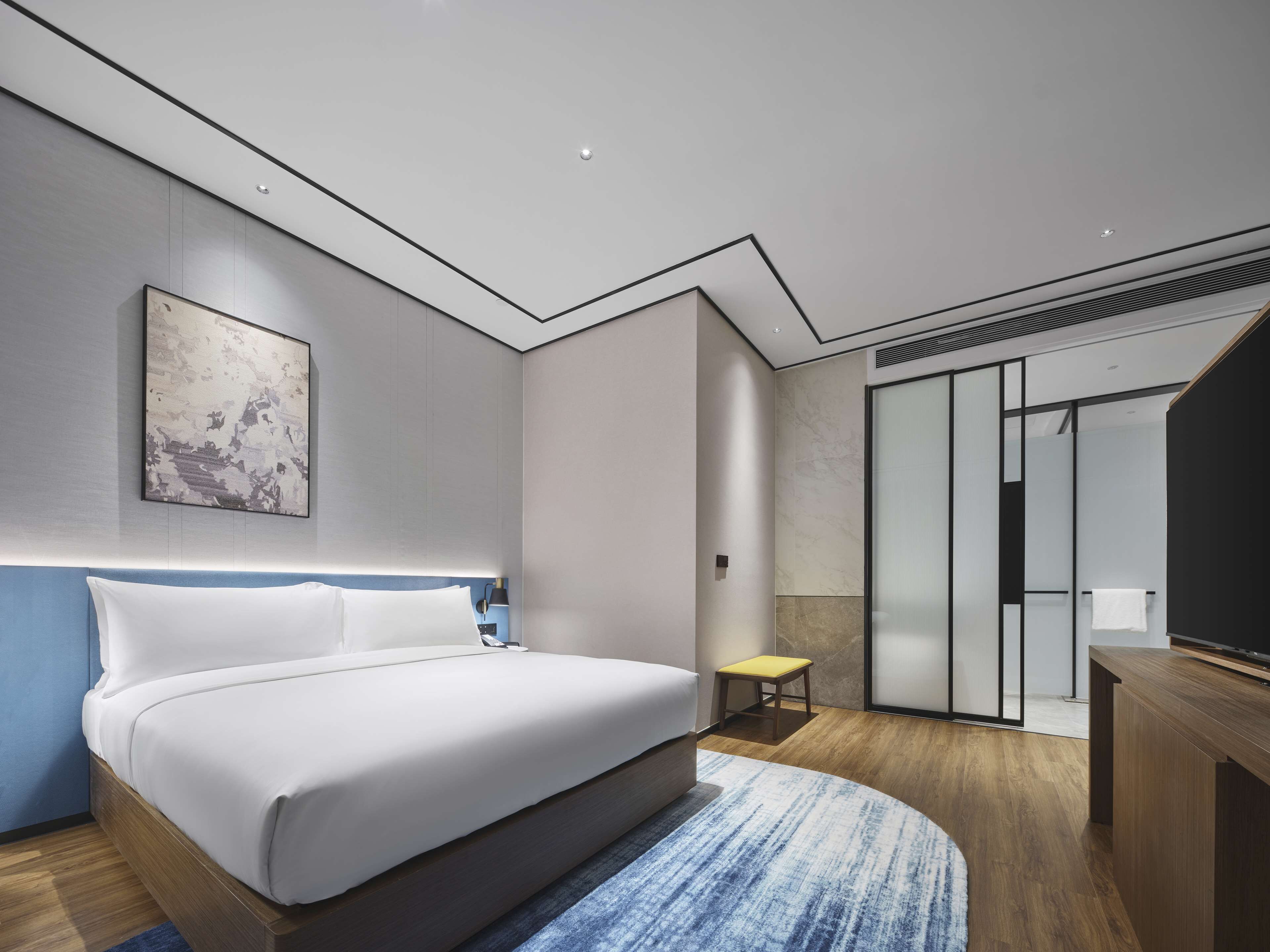 A modern hotel room with a large bed, a small stool, a desk, and a sliding glass door leading to a bathroom.