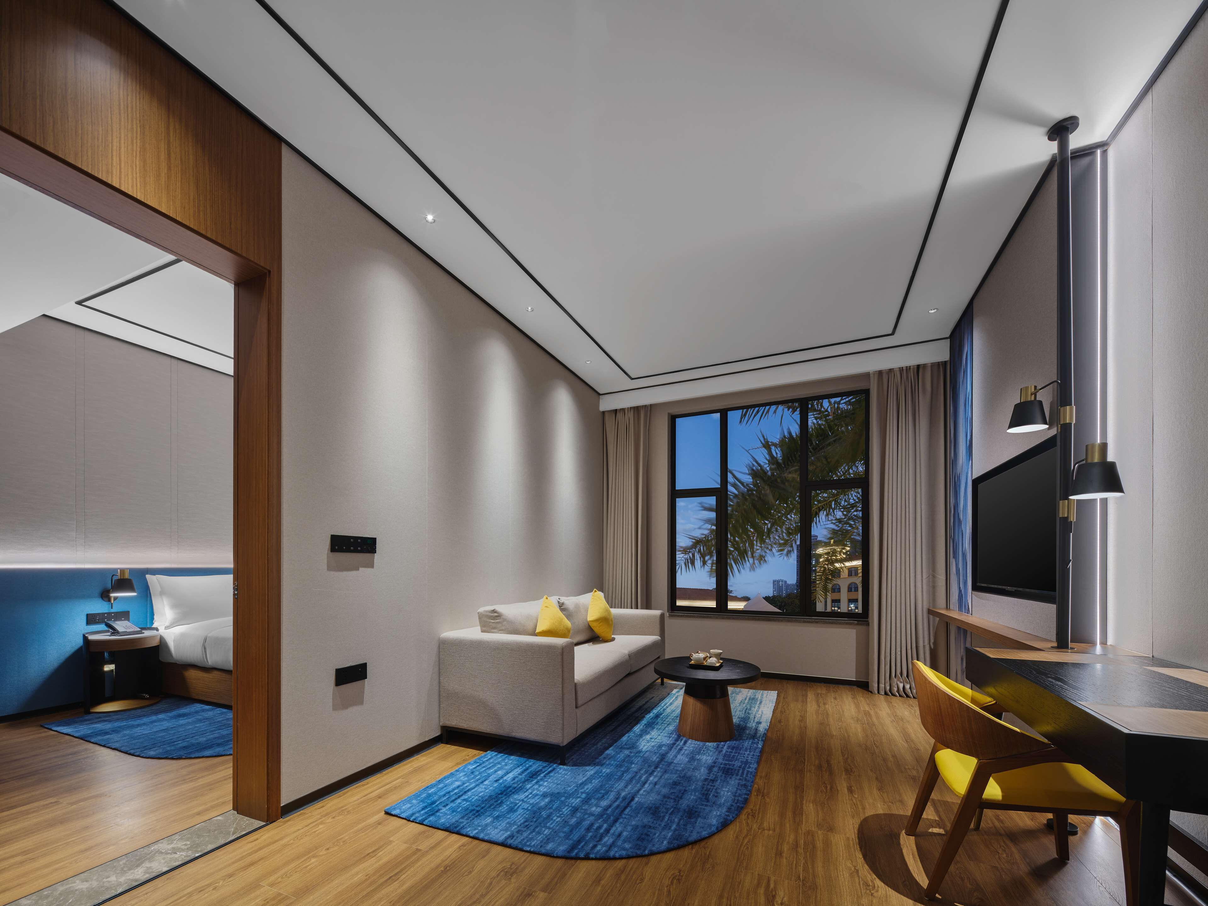 A modern hotel room with a sofa, a dining table with chairs, a TV, and a large window with a view of palm trees.