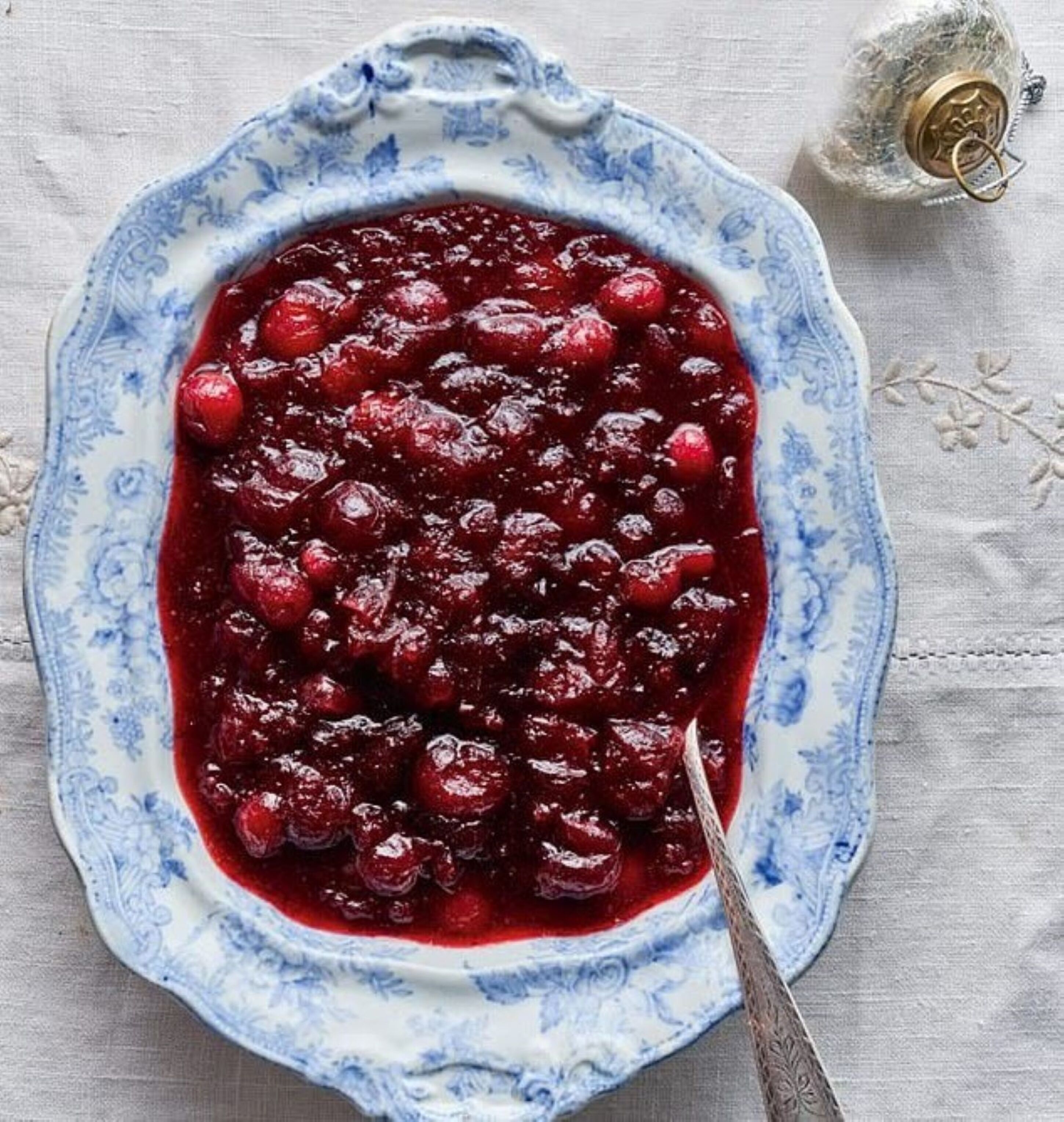 A ceramic dish with a blue floral pattern containing a red fruit compote, accompanied by a spoon and a decorative metal object.