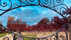 A wrought iron gate with a stone wall and a wooden pathway leading to a historic building with ivy-covered walls and a tower.