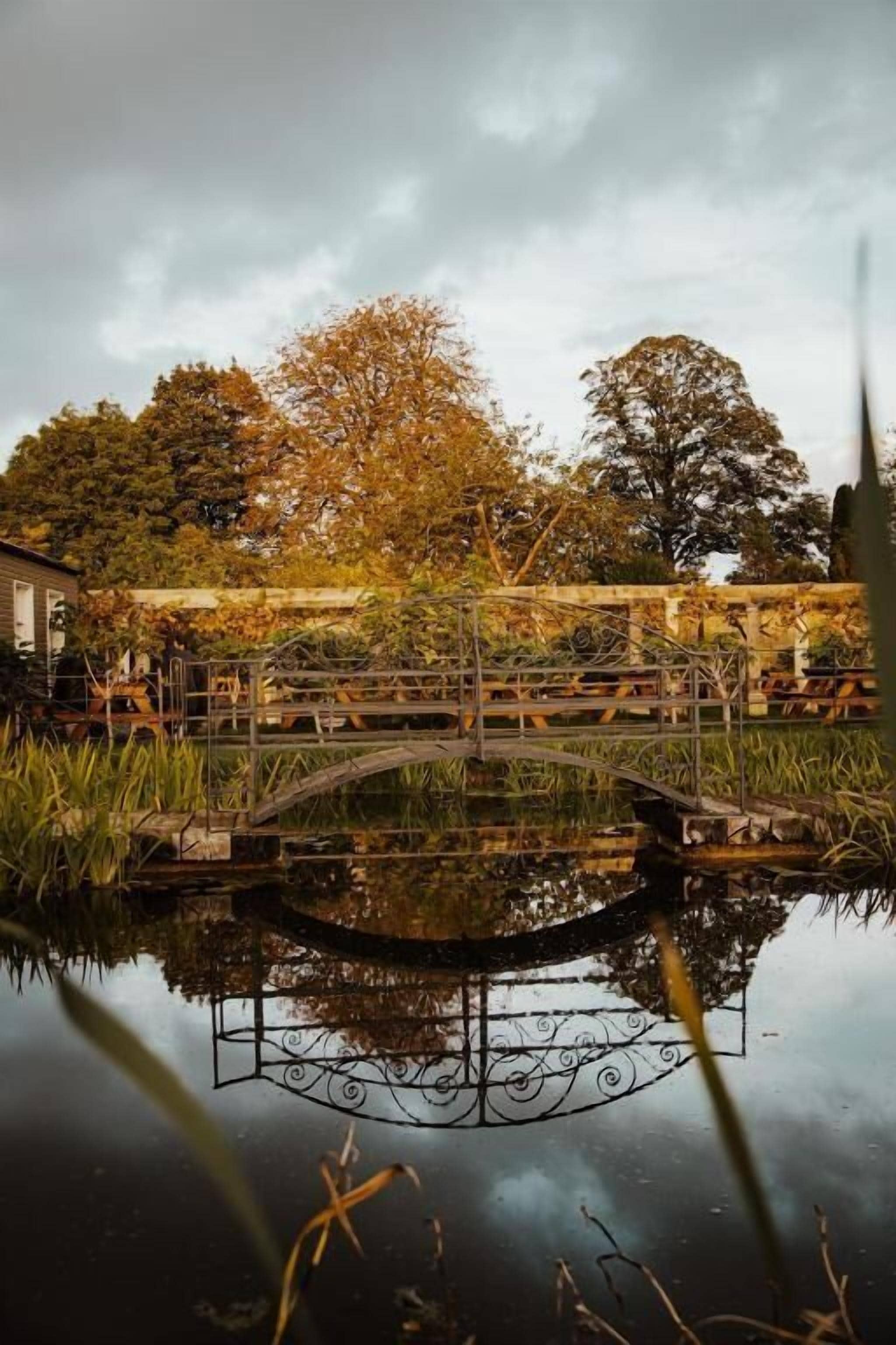 A bridge over a calm water body with a reflection, surrounded by trees and a building in the background.