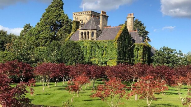 A large, historic building with a prominent tower and multiple chimneys, surrounded by lush greenery and a well-maintained lawn.